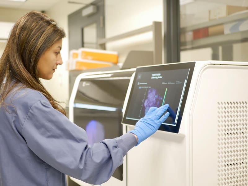 A female scientist in a lab coat and gloves analyzing data on a touchscreen monitor in a laboratory.